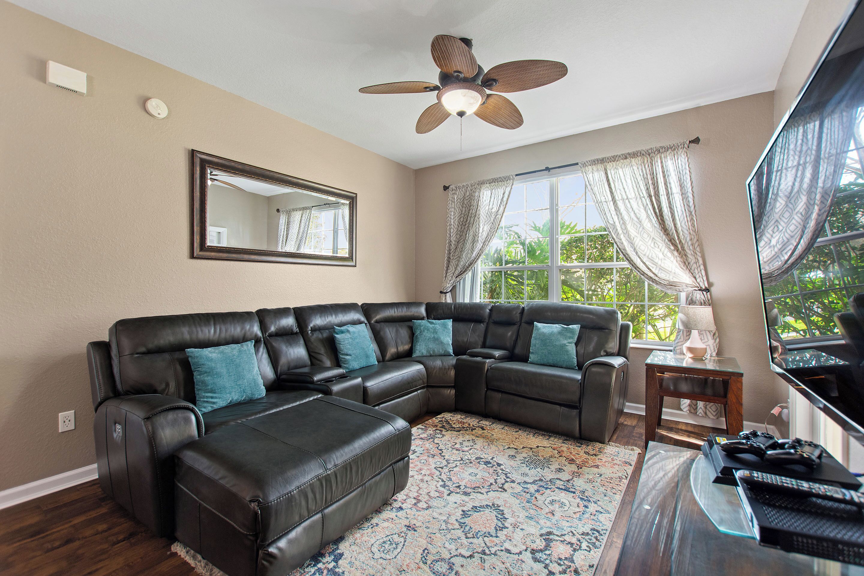 A living room with a sectional sofa, a ceiling fan, a large window with curtains, and a flat-screen TV.