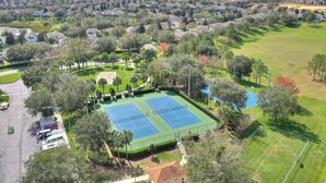 An aerial view of a residential area with two tennis courts surrounded by trees and a parking lot.