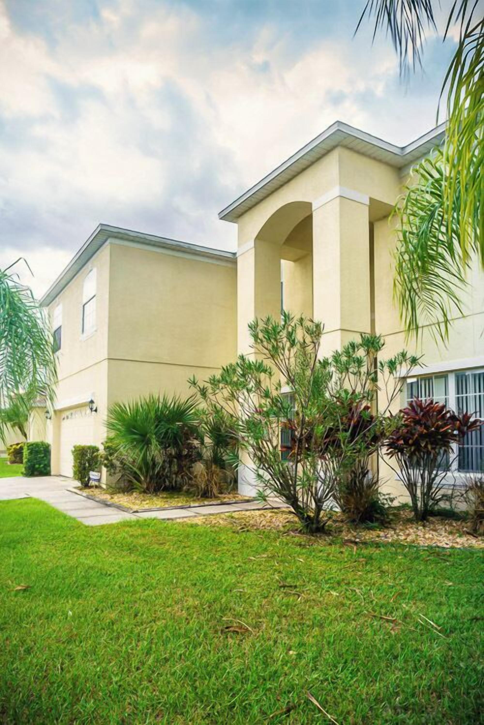 A two-story building with a flat roof and a beige facade. There are small shrubs and palm trees in the front yard.