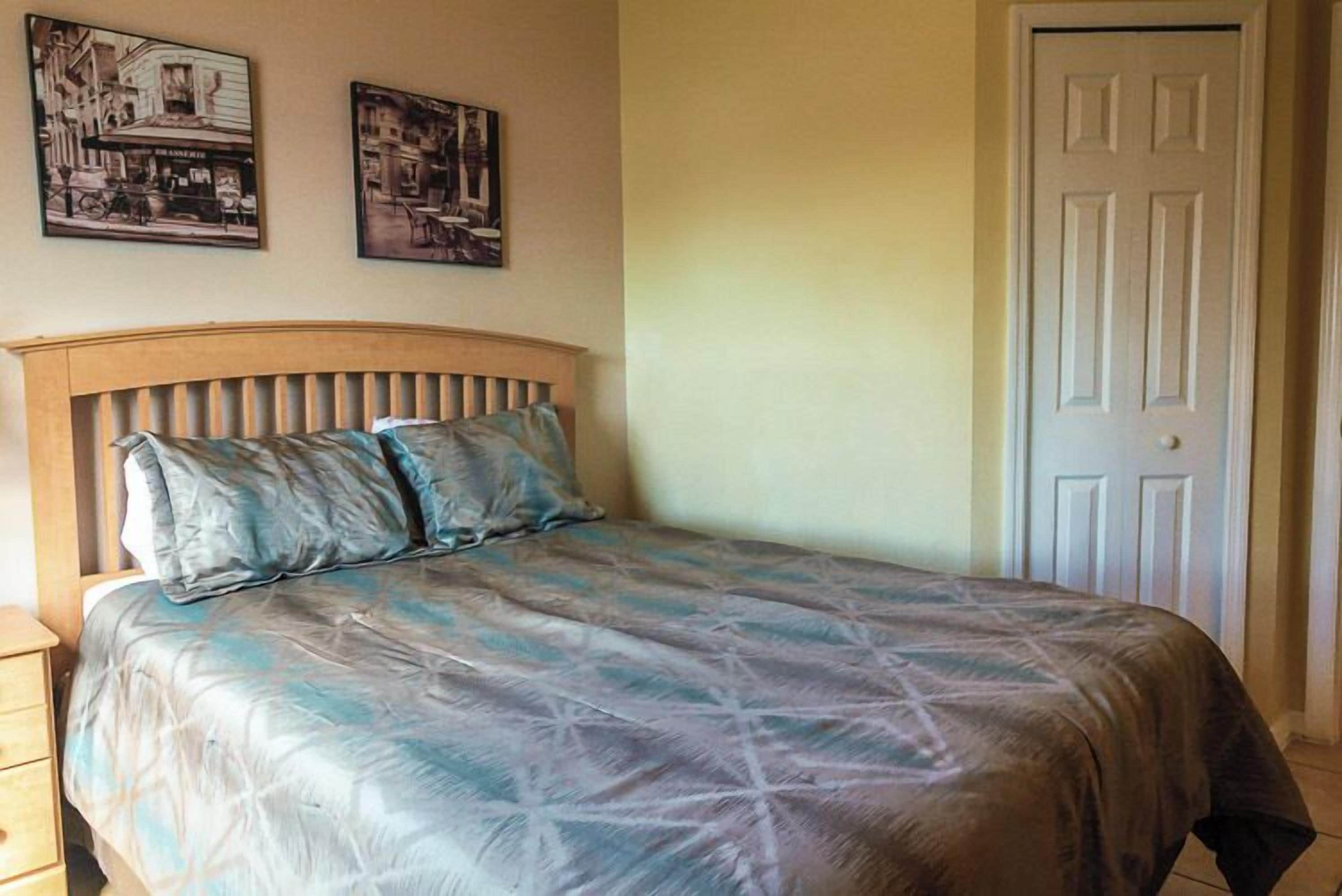 A bedroom with a wooden headboard, a bed with a patterned bedspread, two wall-mounted pictures, and a closed door.