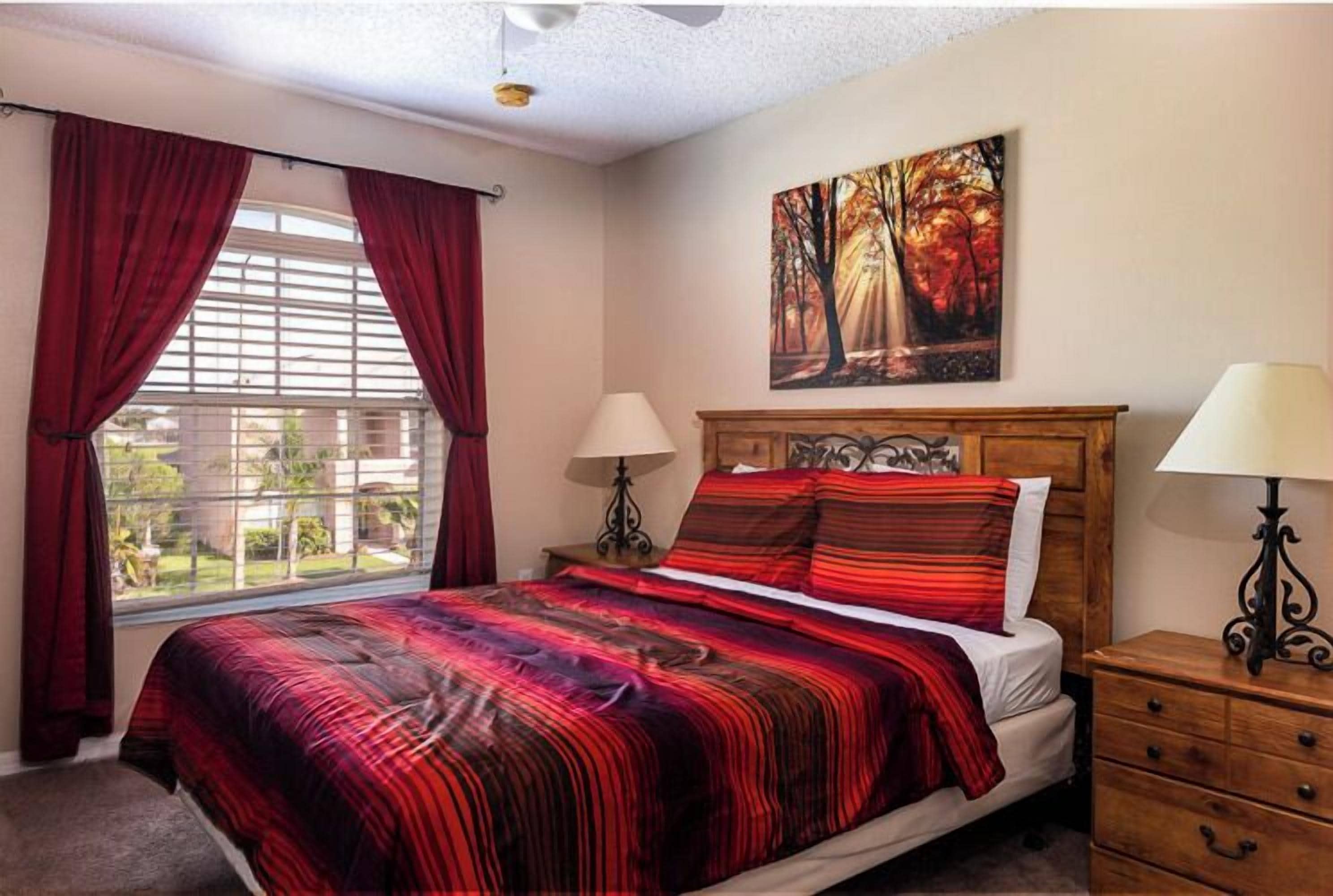 Una camera da letto con un letto grande, tende rosse, un quadro raffigurante alberi autunnali e un comodino in legno con una lampada.
