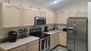 A kitchen with light-colored cabinets, a stainless steel refrigerator, a microwave, a stove, and a coffee maker. - 8054 Hidden Gem (Kissimmee)