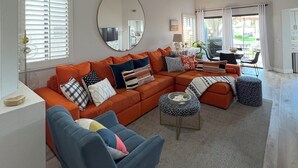 A spacious living room with a large orange sectional sofa, a round mirror, and a ceiling fan. - Stylish 2-bedroom condo in Palm Desert with WiFi, AC, and a golf course! (Palm Desert)