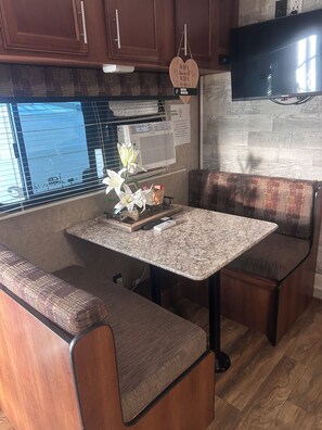 Een compact camperinterieur met een kitchenette, een eethoek met tafel en een bank en een wandgemonteerde flatscreen-tv.