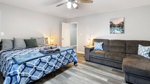 A bedroom with a bed, a gray couch, a ceiling fan, and a door leading to another room.