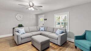 A living room with a sectional sofa, an ottoman, a teal armchair, a ceiling fan, and a clock on the wall.