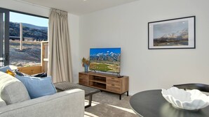 A modern living room with a flat-screen TV, a wooden entertainment unit, a grey sofa, a dining table, and a view of a mountainous landscape through large windows. - Peaceful, Luxury Unit At The Iconic Remarkables! (Queenstown)