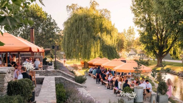 An outdoor patio area with stone steps leading to tables under orange umbrellas, surrounded by greenery and a pond. - Peaceful, Luxury Unit At The Iconic Remarkables! (Queenstown)