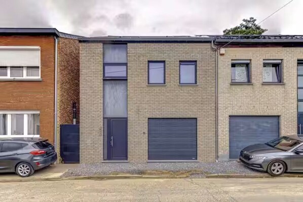 Une rangée d’immeubles à trois étages, avec une porte de garage sur le côté droit, une voiture garée devant et un ciel nuageux.