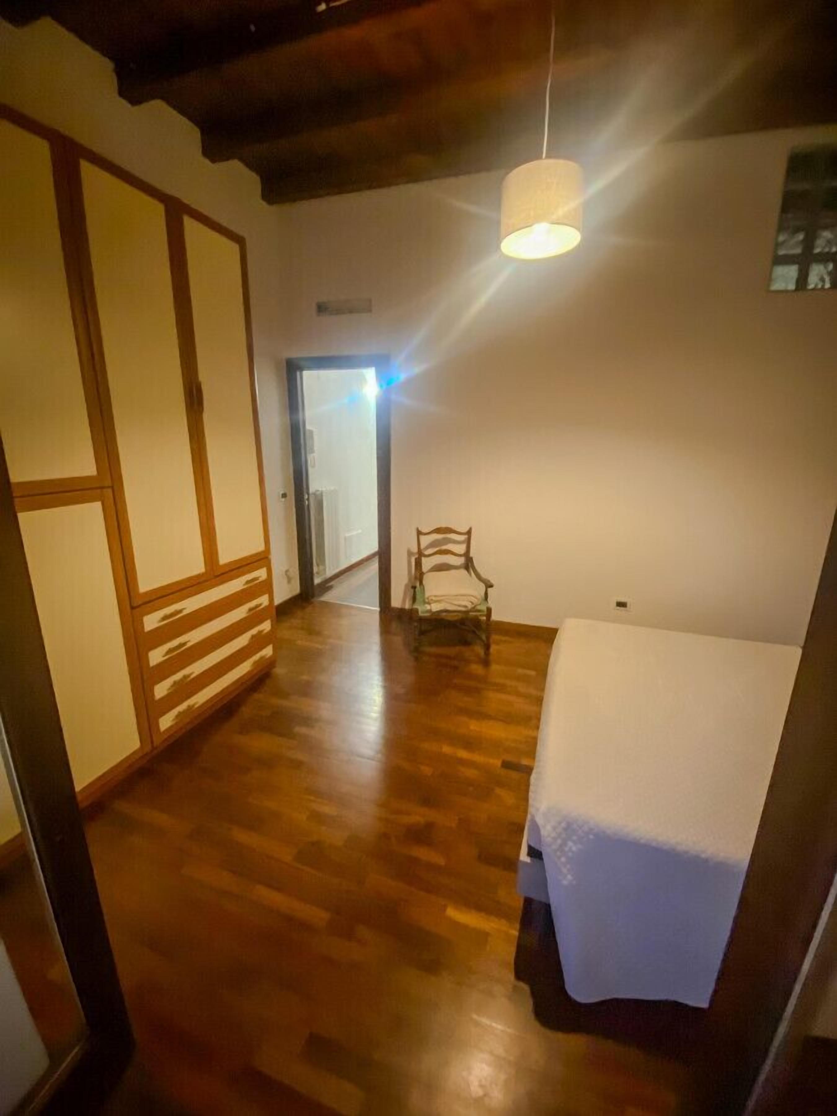 Una habitación con piso de madera, una silla, una cama con cubierta blanca y una lámpara colgante.