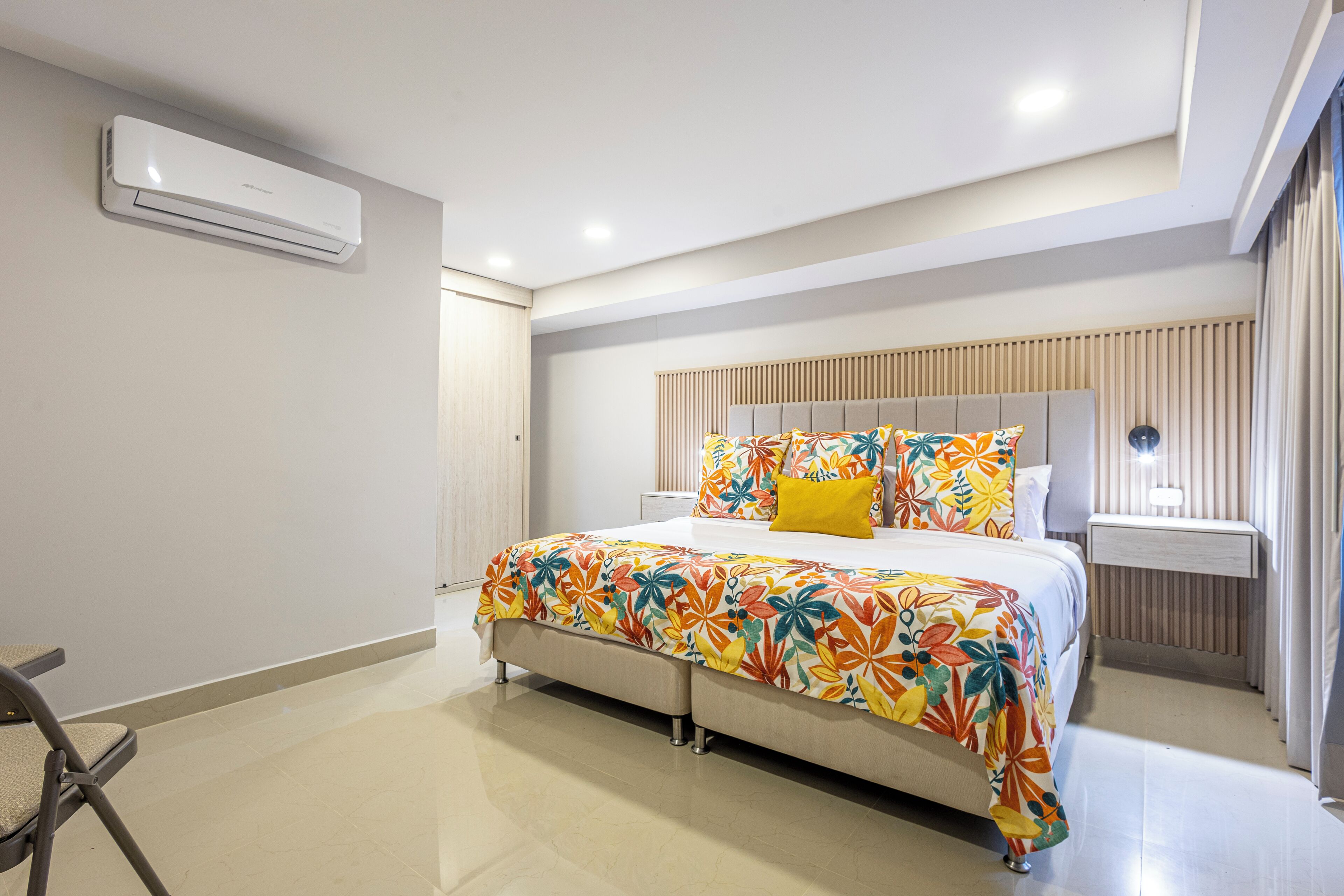 A modern hotel room with a large bed, a bedside table, a chair, and an air conditioning unit on the wall.