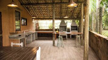 Uma área de cozinha externa com mesa de madeira, banquetas de bar e churrasqueira.