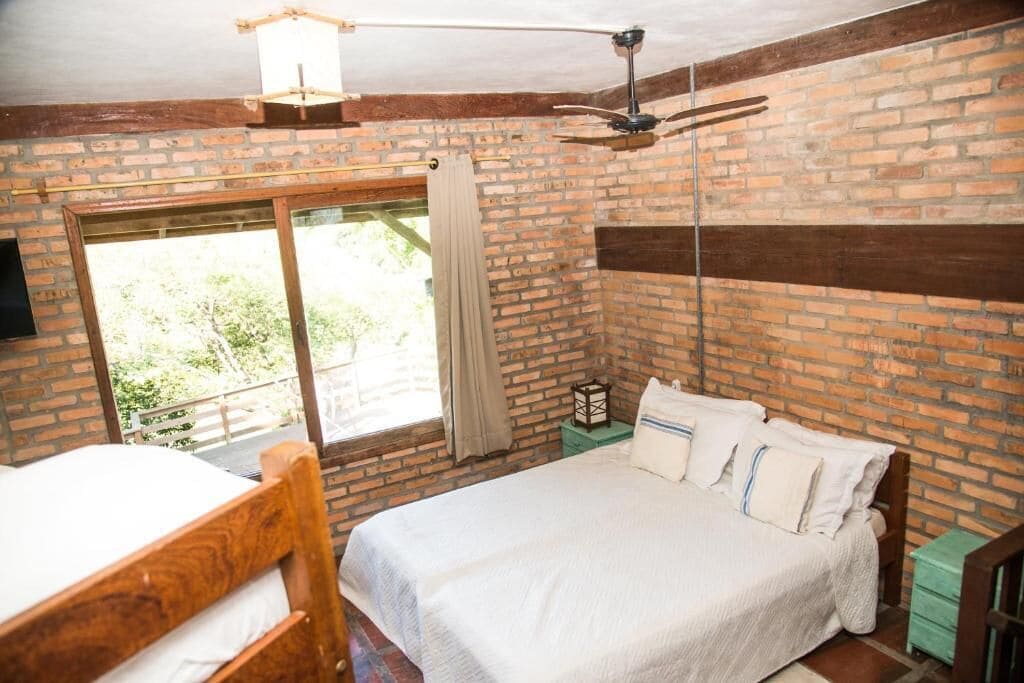 Un dormitorio con pared de ladrillo, cama de madera, ventana con cortinas y ventilador de techo.