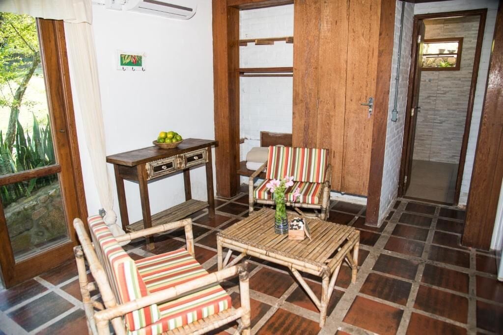 Una habitación con piso de madera, una mesita con un florero, dos sillas a rayas, un banco de madera y una puerta que da a otra habitación.