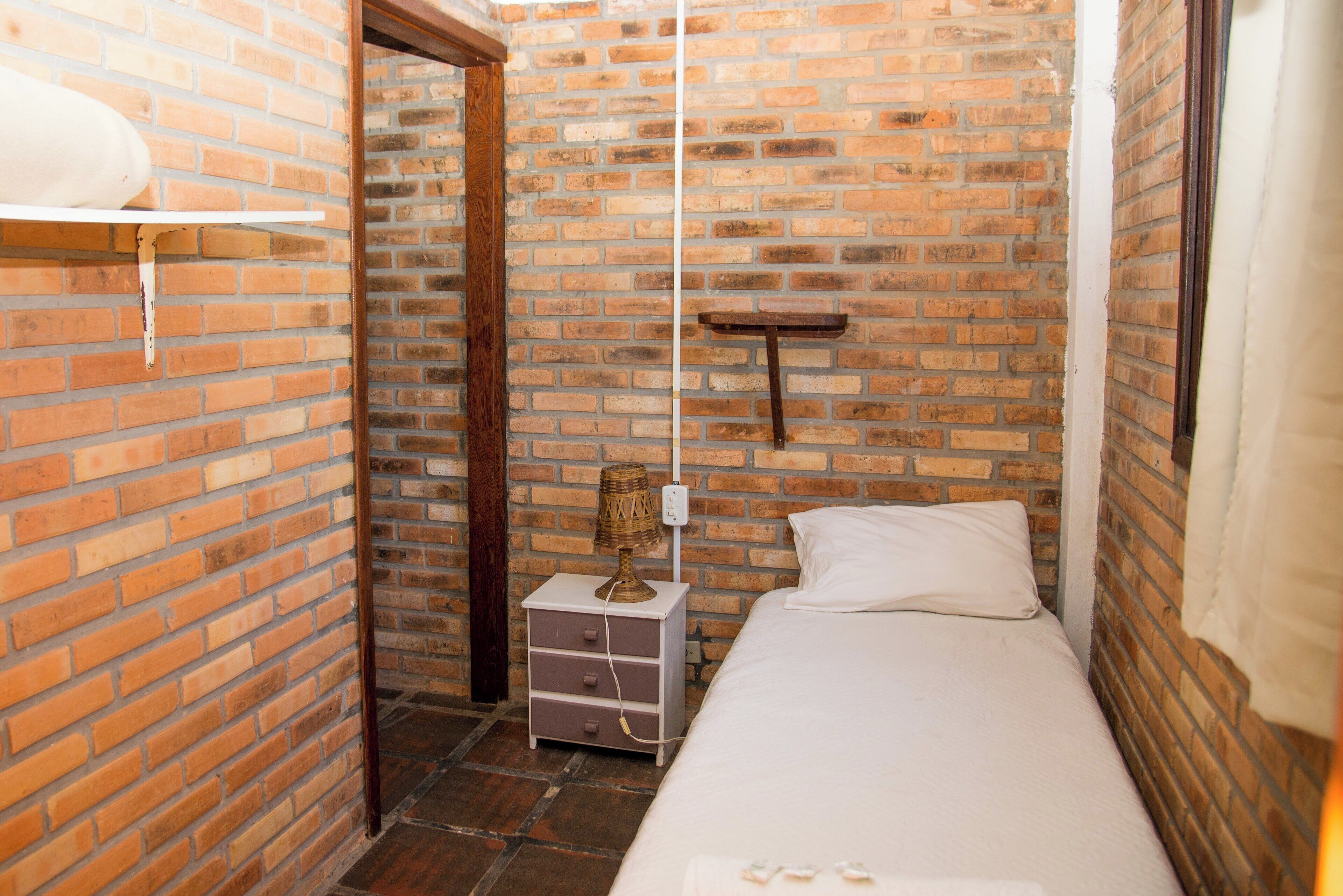 Una habitación estrecha con pared de ladrillo, cama, mesita de noche y estante.