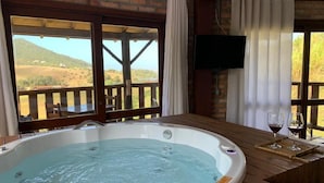 A hot tub in a room with a view of a hillside, a wooden deck, and a mounted TV. - Pousada La Creacion (Imbituba)