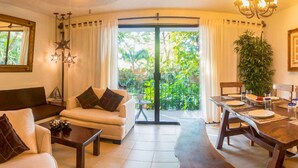 A living room with a sofa, coffee table, and dining table set for a meal. A large window with curtains and a view of greenery is visible. - Cozy stay Tropical Vibes near Beach (Puerto Vallarta)