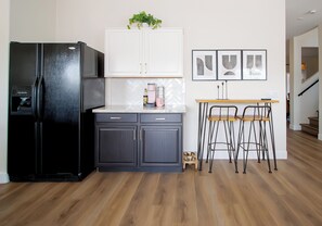 Een keuken met een zwarte koelkast, donkere kastjes, een wit aanrecht, een klein houten tafeltje met krukken en lijstwerken aan de muur.