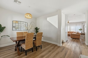 Un comedor con una mesa de madera y sillas tejidas, una lámpara colgante y un espejo con diseño de cuadrícula en la pared.