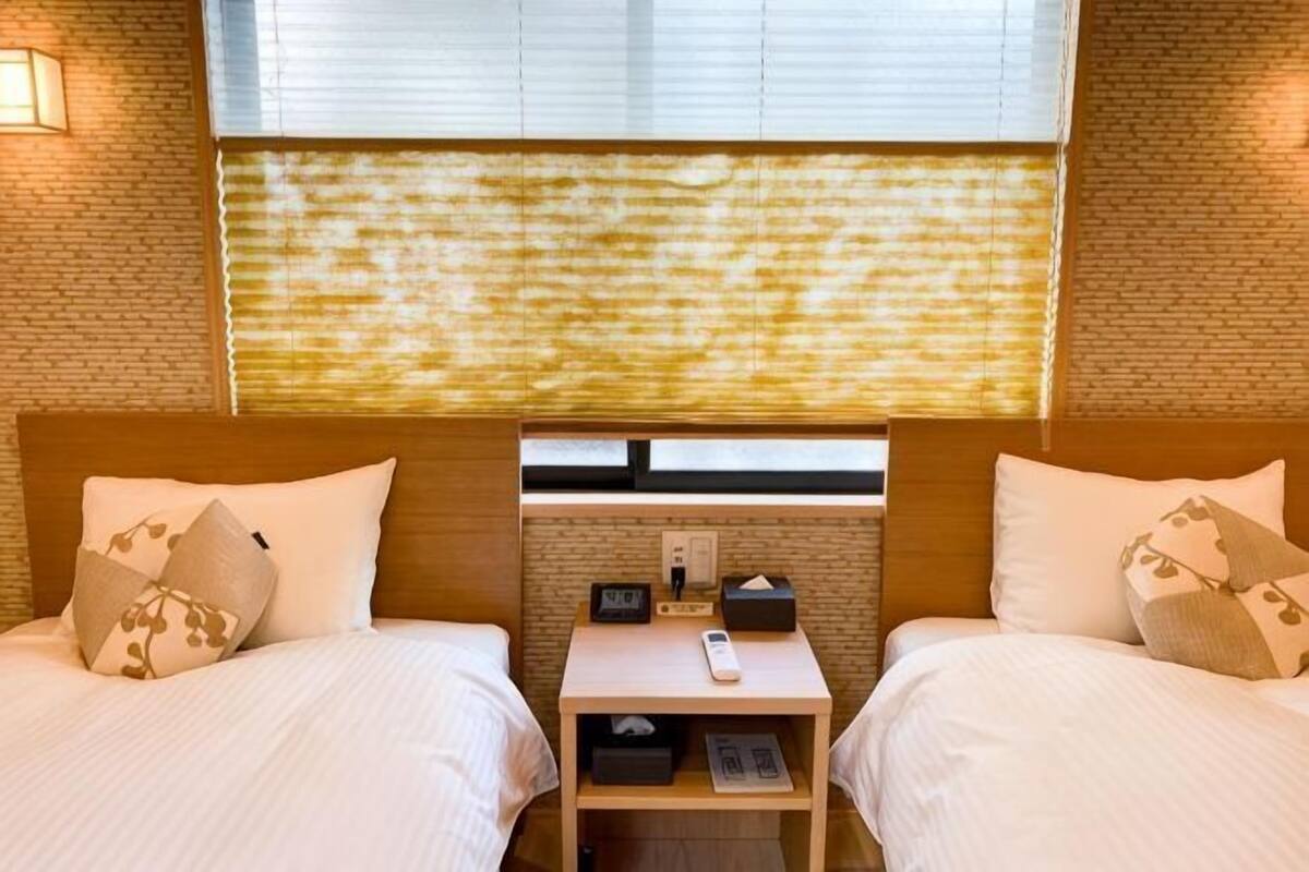A hotel room with two beds, a nightstand, a window with blinds, and a brick wall.