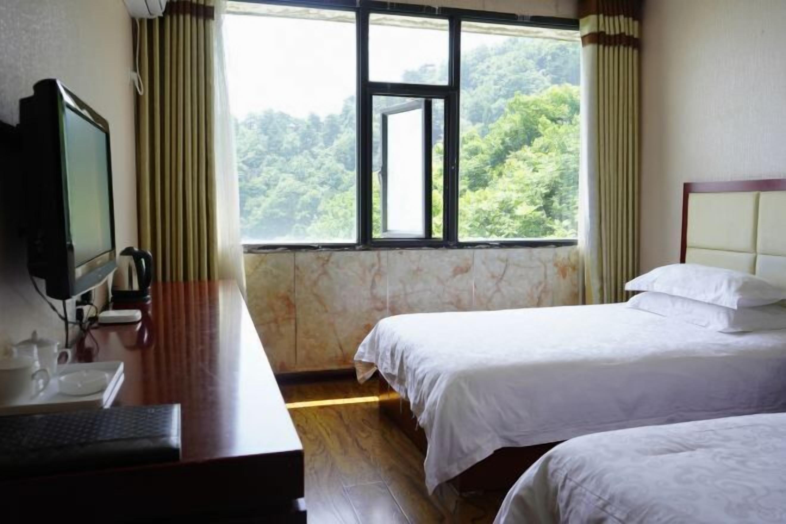 A hotel room with a bed, a desk with a computer, a TV, and a window with a view of a forest.
