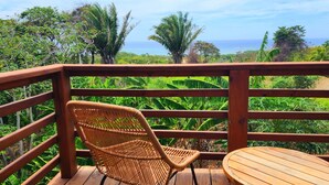 A wooden deck with a wicker chair and a small round table, overlooking a lush landscape with dense vegetation and a view of the ocean. - Stunning Ocean Views, Peaceful, Private Pool, Lush Natural Surroundings (Roatan)