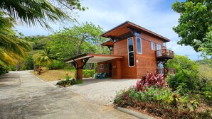 A wooden house with a covered porch, surrounded by lush greenery and a paved driveway. - Stunning Ocean Views, Peaceful, Private Pool, Lush Natural Surroundings (Roatan)