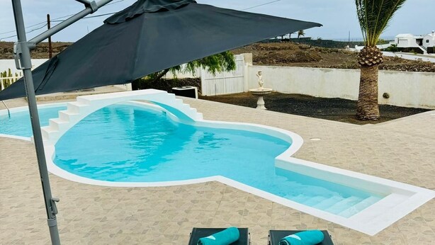 A swimming pool with a unique shape, surrounded by a tiled patio. There are two lounge chairs with towels, a large umbrella, and a palm tree in the background. - Ferienwohnung in Einer Finca 4000m2 mit Pool Naehe Puerto Calero (Mácher)