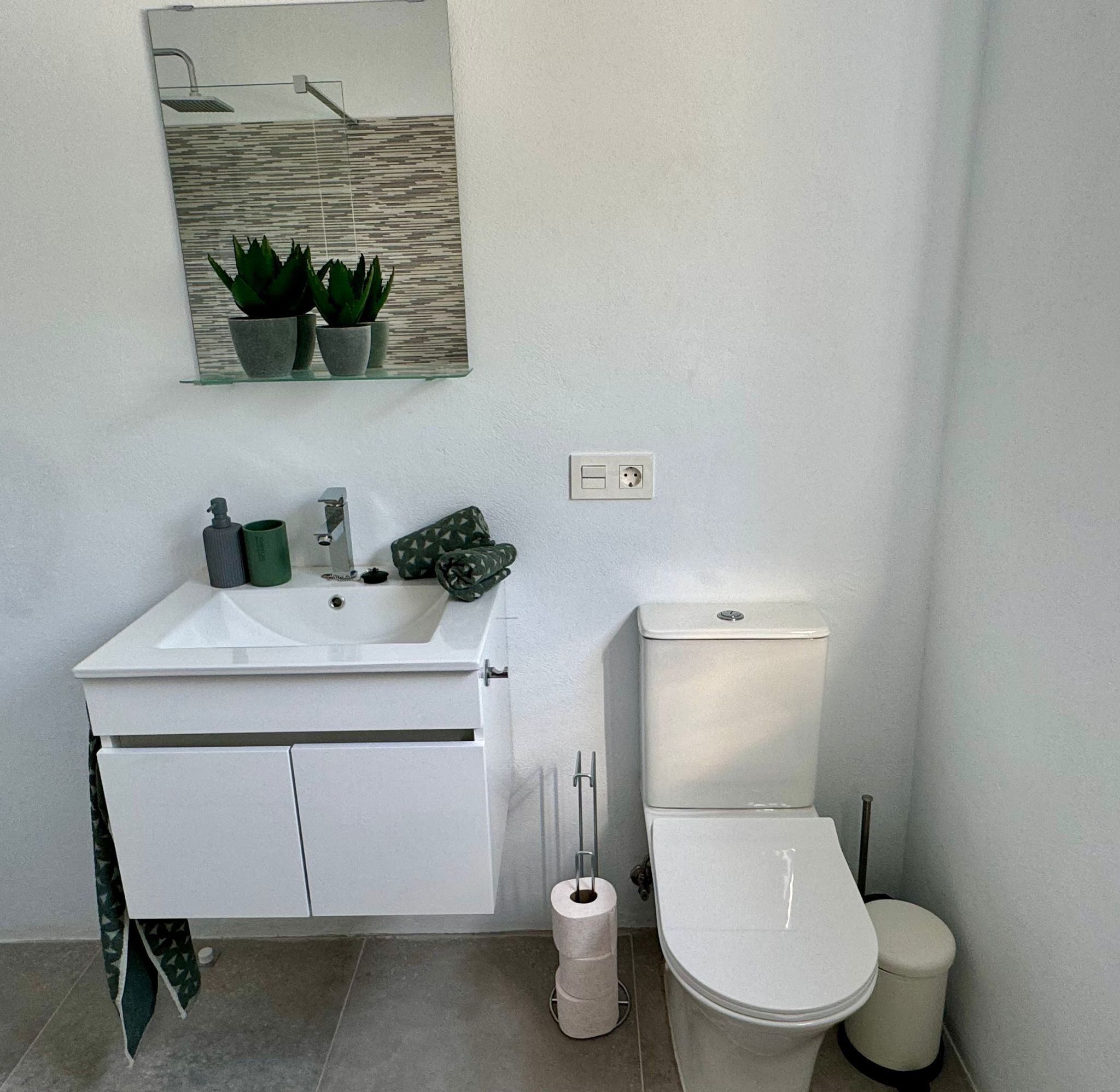 A bathroom with a white sink, mirror, toilet, and a small trash bin.