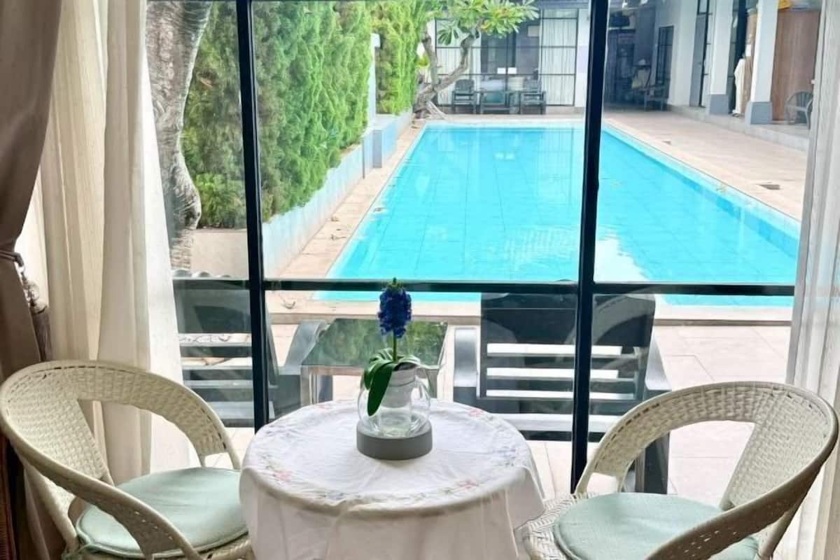 Une chambre avec vue sur une piscine extérieure, des fauteuils en osier, une petite table avec un vase et une fenêtre avec des rideaux.