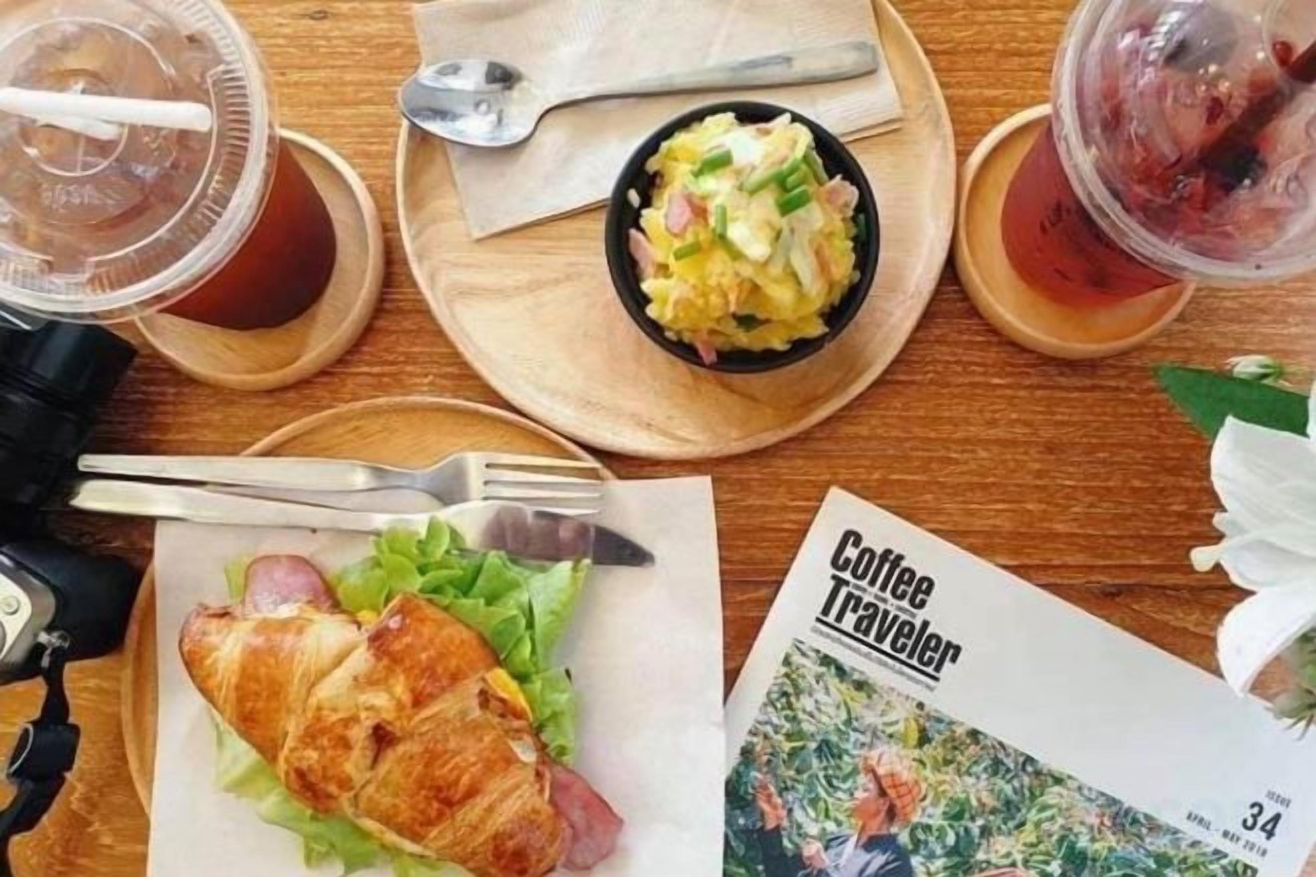 Ateria, johon kuuluu croissant, salaatti ja juomia, ja se nautitaan puupöydän ääressä lehden parissa.