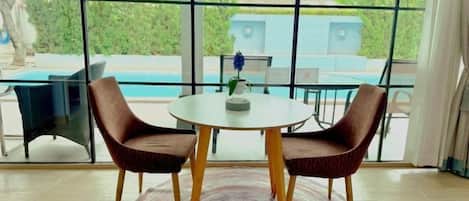 Una habitación con una mesa redonda y dos sillas, vista a una piscina a través de amplios ventanales, y un entorno tropical al aire libre.