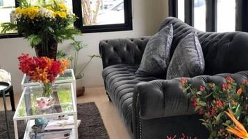 Un salón con un sofá de color oscuro, una mesa de centro de vidrio con revistas y un jarrón con flores.