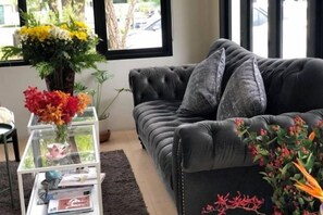 Un salon avec un canapé de couleur sombre, une table basse en verre sur laquelle sont posés des magazines, et un vase rempli de fleurs.