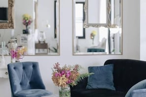 A room with a dark-colored sofa, a blue armchair, and a vase with flowers. There are mirrors on the wall and a white shelf with items on it. - La Riva Boutique Hotel (Kamphaeng Phet)