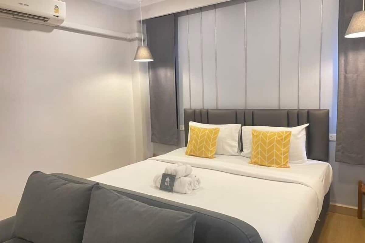 Une chambre d’hôtel avec un lit, un canapé gris, un climatiseur fixé au mur et un luminaire suspendu.