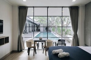 A hotel room with a bed, a desk, a chair, a TV, and a view of an outdoor swimming pool.