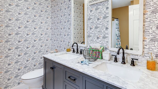 Une salle de bain avec un papier peint à motifs de poissons, des toilettes blanches, un lavabo avec un plan de travail en marbre et un miroir avec une plaque nominative.