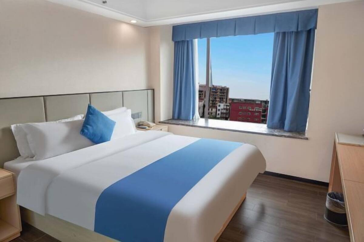 A hotel room with a large bed, a nightstand, a window with curtains, and a view of buildings.
