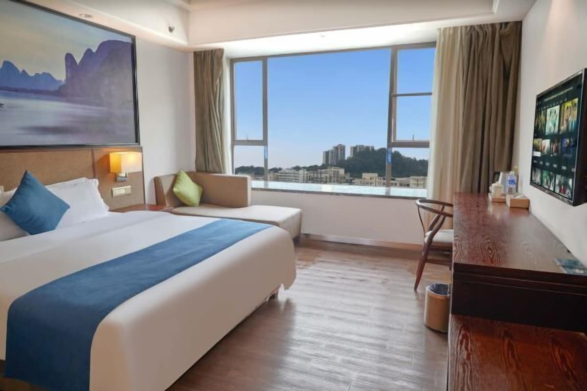 Ein Hotelzimmer mit einem großen Bett, einem Schreibtisch, einem Sessel, einem Fernseher und einem Fenster mit Blick auf die Stadt.