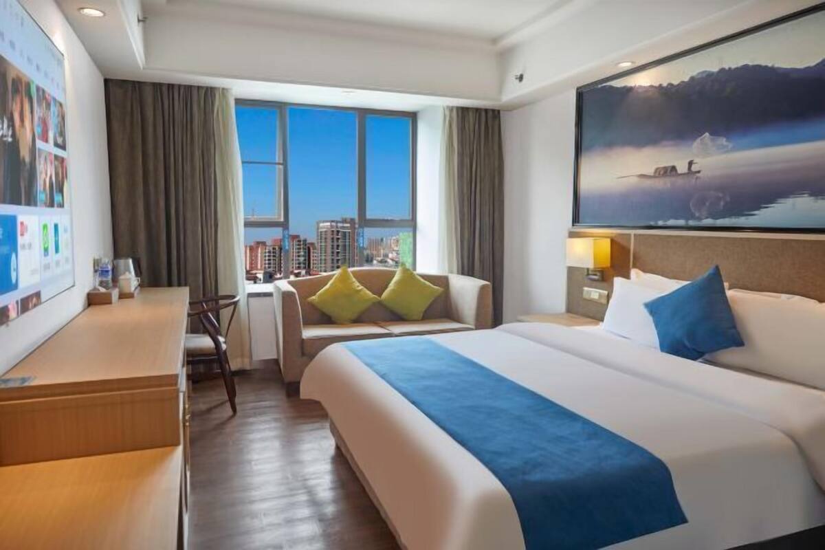 A hotel room with a large bed, a desk, a sofa, and a view of the cityscape through the window.