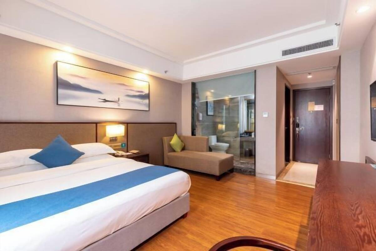 A hotel room with a bed, a sofa, a desk, a TV, and a bathroom visible through a glass partition.
