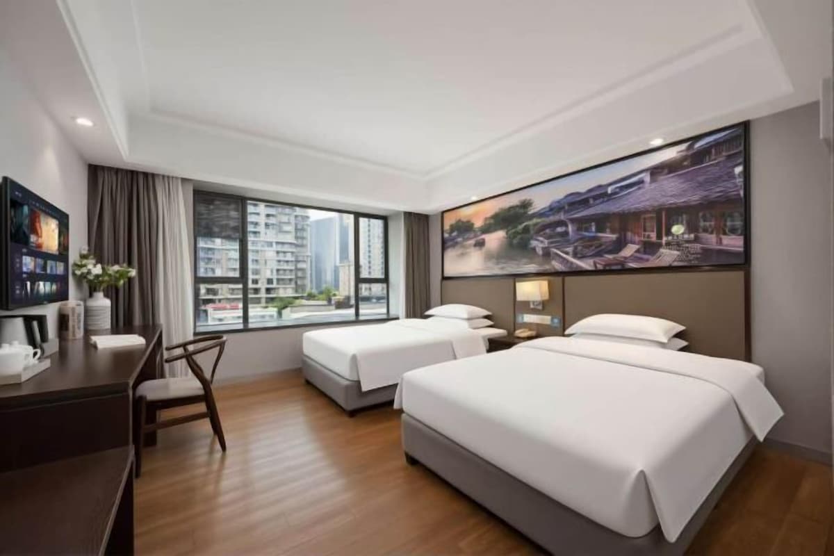 A hotel room with two beds, a desk, a chair, a TV, and a large window with a city view.