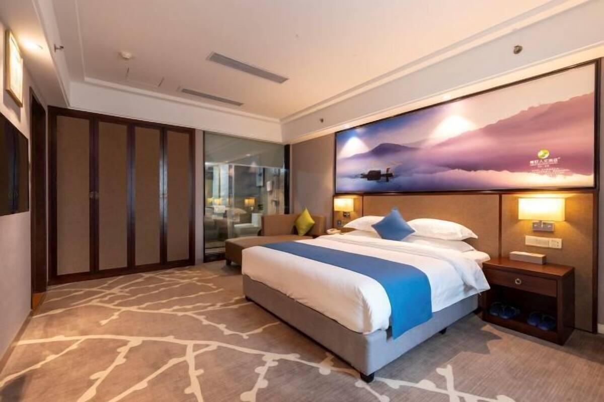 A hotel room with a bed, a nightstand, a wall-mounted TV, and a large window.