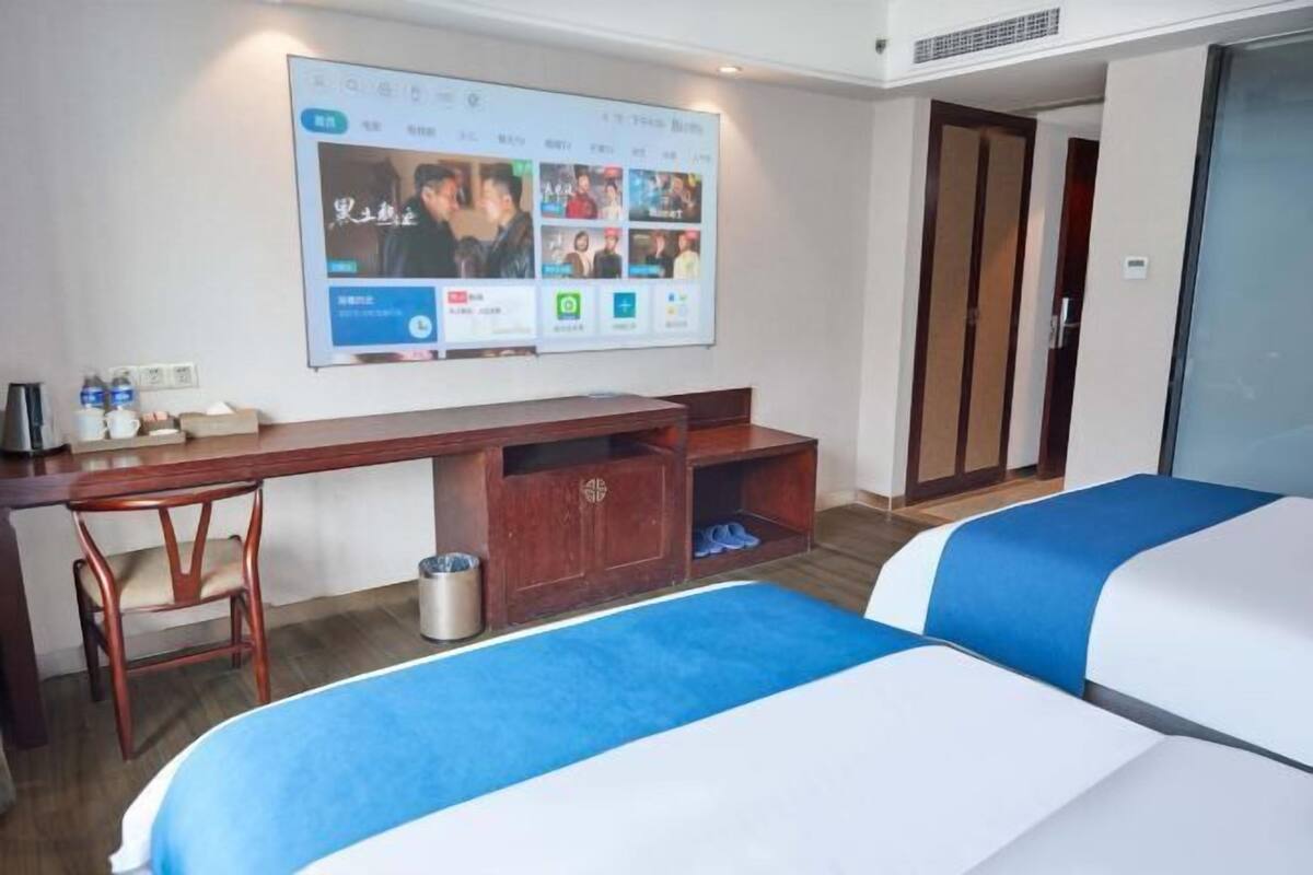 A hotel room with a desk, two beds, a TV screen, and a shoe rack.