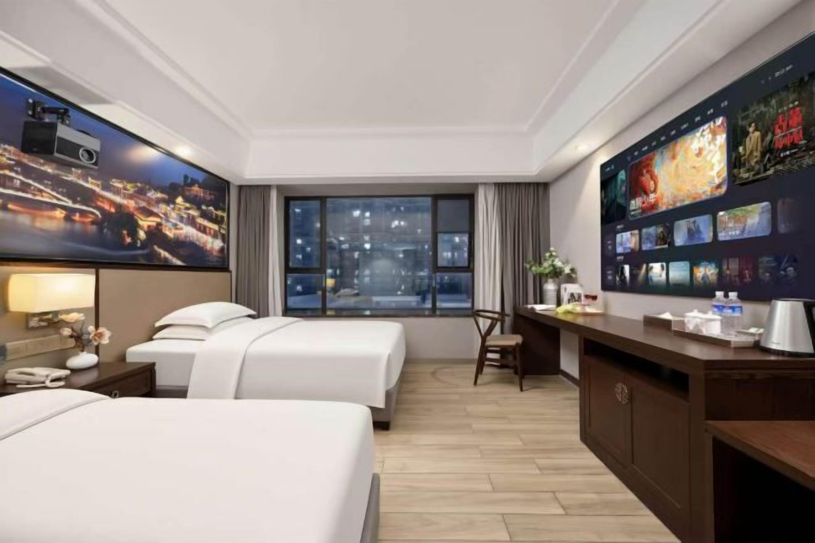 A hotel room with two beds, a desk, a TV wall, and a large window with a city view.