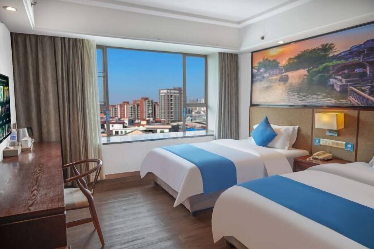 A hotel room with two beds, a desk, a chair, a painting, and a view of a cityscape.