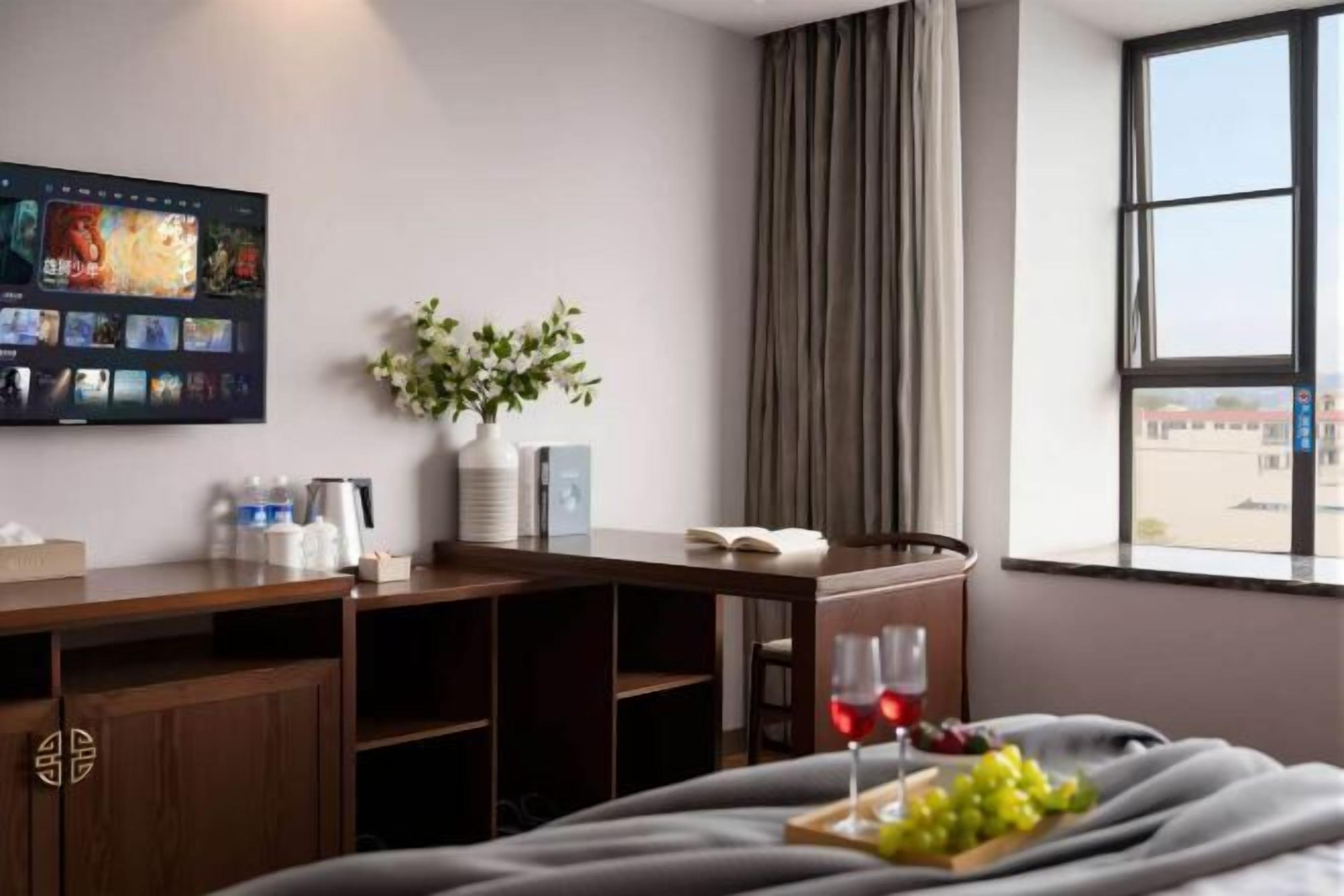 Ett hotellrum med en säng, ett skrivbord med en bärbar dator, en vas med blommor, ett fönster med gardiner och en tv på väggen.