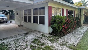 Ett envåningshus med ett garage med tak, en liten trädgård med röda blommor och en palm i bakgrunden.