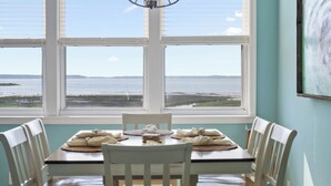 Ein Esstisch mit weißen Stühlen und Blick auf das Meer durch große Fenster.